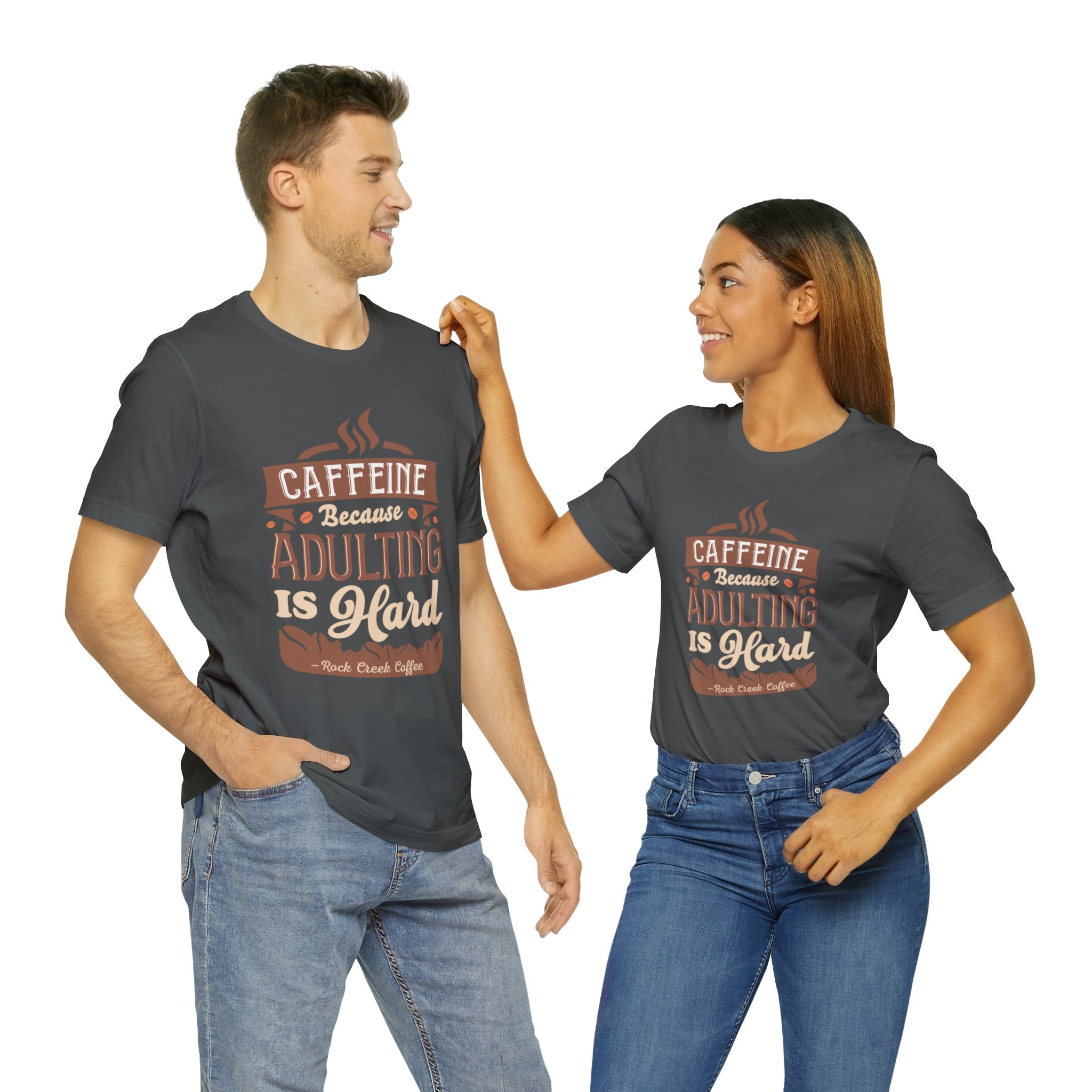 Adult man and woman modeling matching asphalt short-sleeve T-shirts featuring the “Caffeine Because Adulting Is Hard” graphic in a stacked typographic design, posed together against a plain white background.