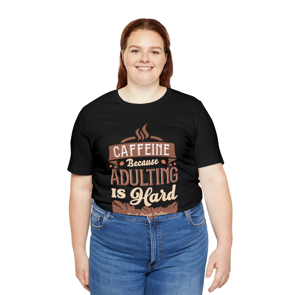 Adult woman smiling while modeling a black short-sleeve T-shirt featuring the “Caffeine Because Adulting Is Hard” graphic in a stacked typographic design, shown against a plain white background.