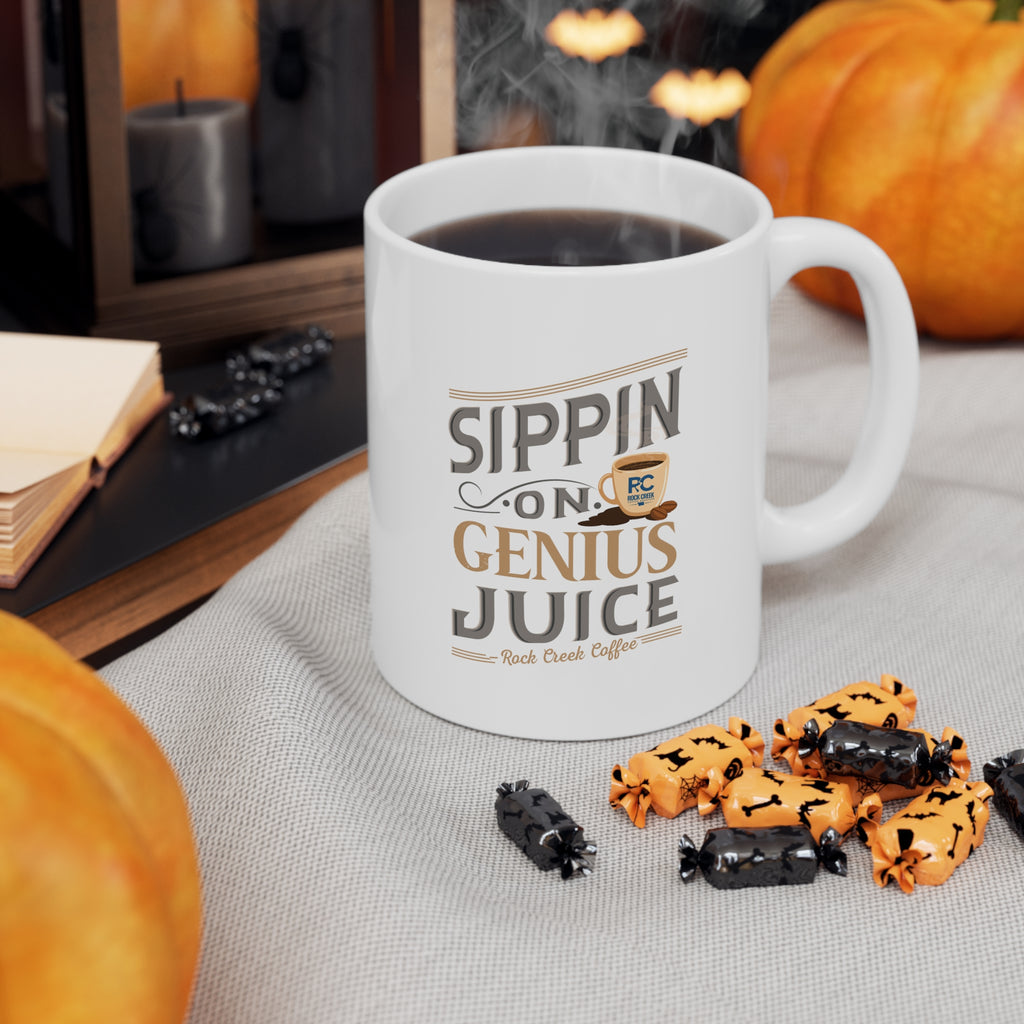 White Rock Creek Coffee mug featuring the phrase “Sippin on Genius Juice” in stylized gray and gold lettering with a small coffee cup graphic, filled with coffee and styled with pumpkins and Halloween candy on a cozy fall themed tabletop.