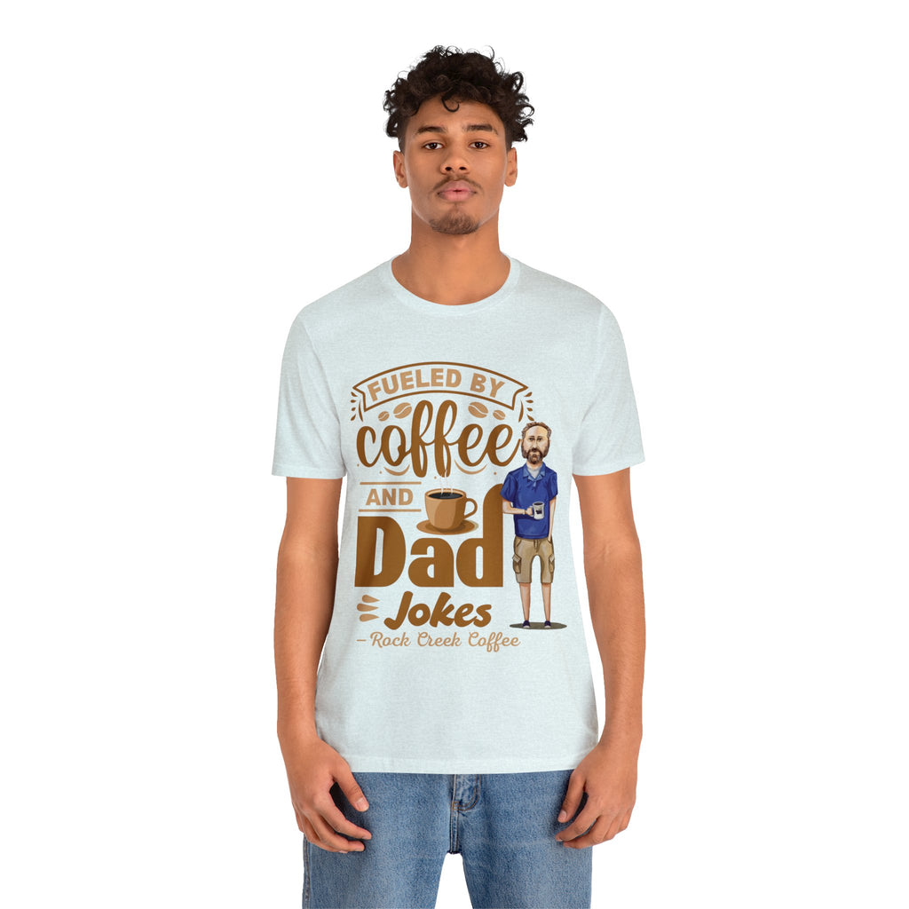 Young man modeling a light blue heather short-sleeve T-shirt with a coffee-themed printed graphic that reads “Fueled by Coffee and Dad Jokes” featuring a steaming coffee cup and cartoon dad illustration, shown against a plain white background.