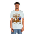 Young man modeling a light blue heather short-sleeve T-shirt with a coffee-themed printed graphic that reads “Fueled by Coffee and Dad Jokes” featuring a steaming coffee cup and cartoon dad illustration, shown against a plain white background.