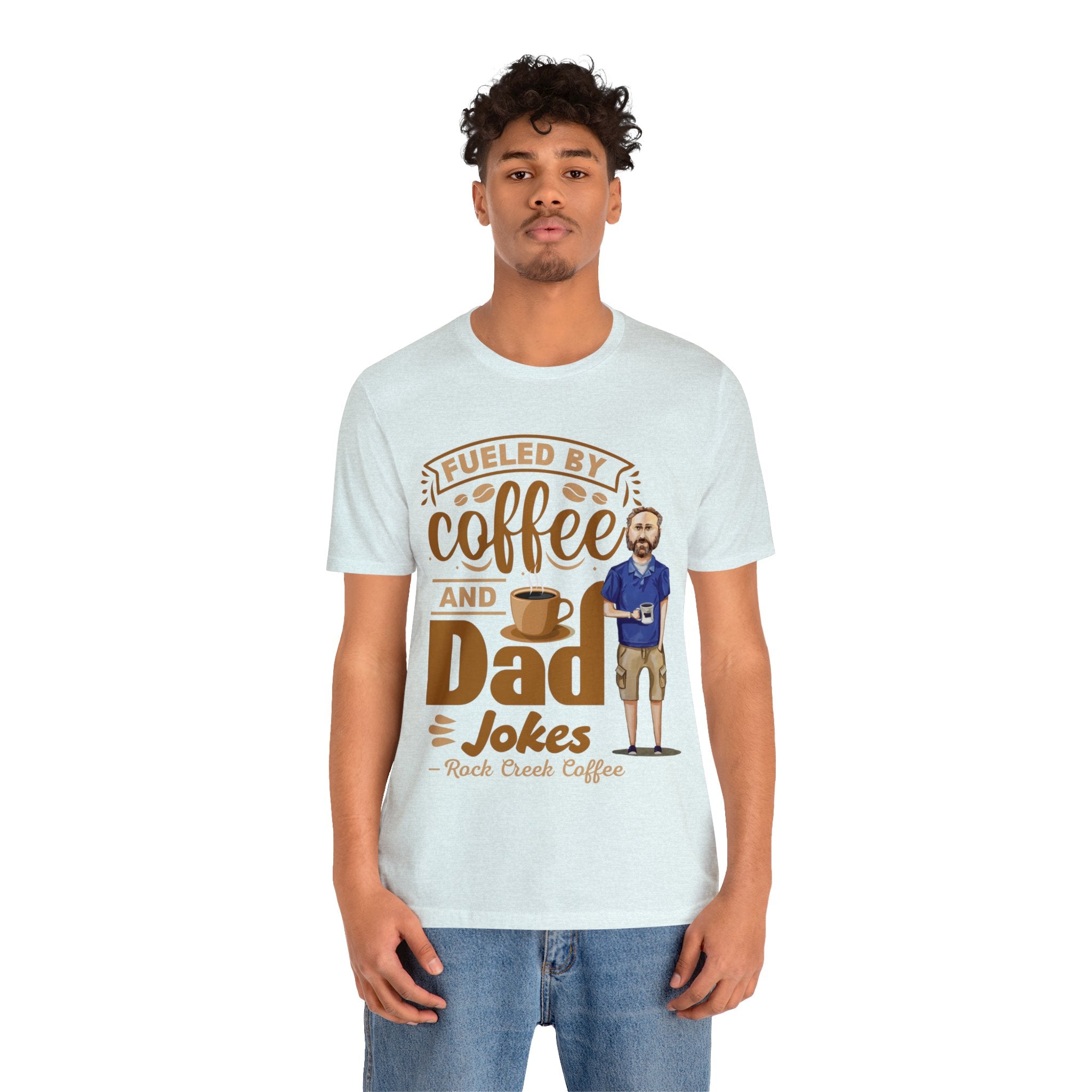 Young man modeling a light blue heather short-sleeve T-shirt with a coffee-themed printed graphic that reads “Fueled by Coffee and Dad Jokes” featuring a steaming coffee cup and cartoon dad illustration, shown against a plain white background.