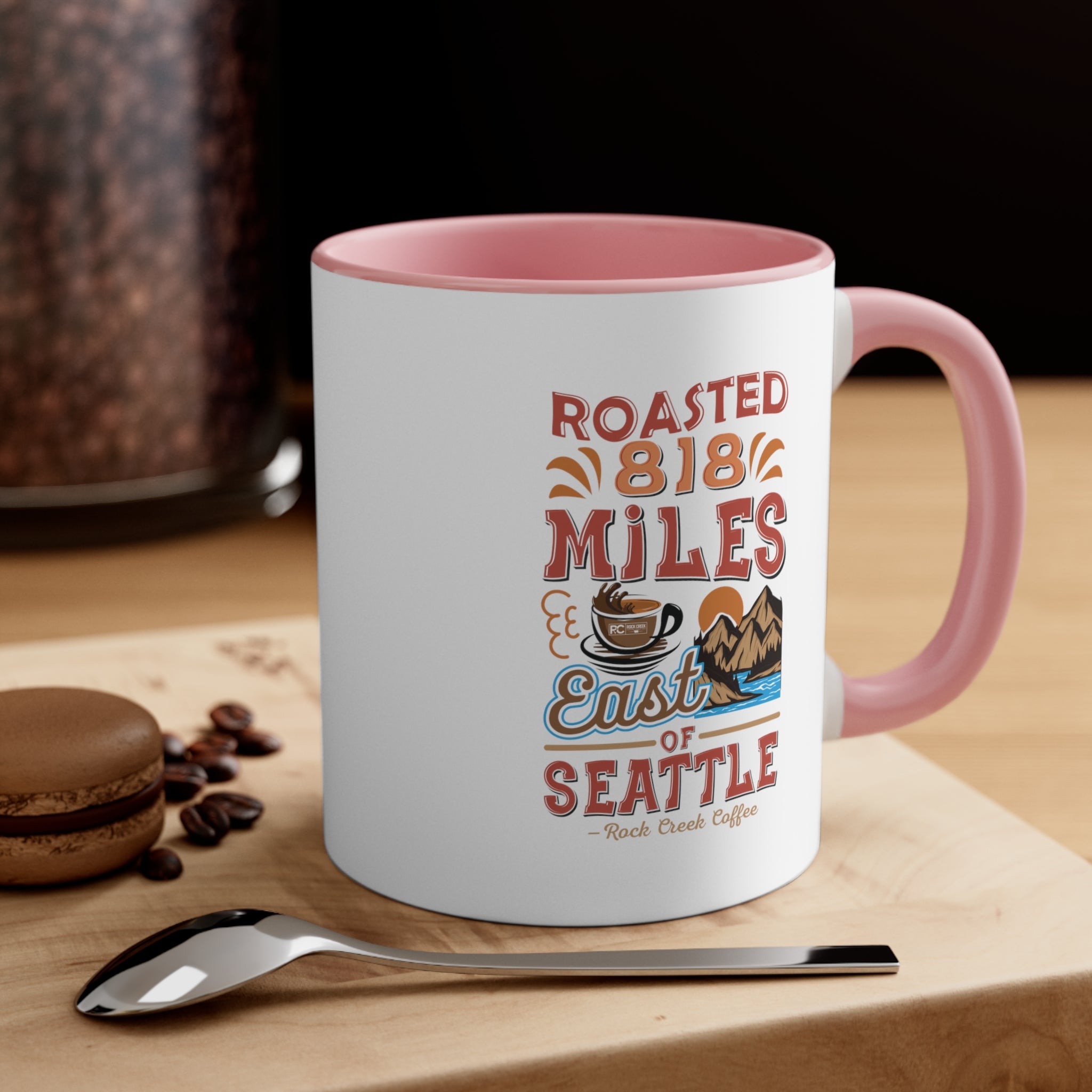 White ceramic coffee mug with pink handle and pink interior featuring the Roasted 818 Miles East of Seattle Rock Creek Coffee design, styled on a wooden surface with a spoon, coffee beans, and a chocolate macaron.