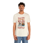 Adult man modeling a cream short-sleeve T-shirt printed with a colorful graphic that reads “Roasted 818 Miles East of Seattle,” featuring a steaming coffee cup and mountain lake illustration, shown against a plain white background.