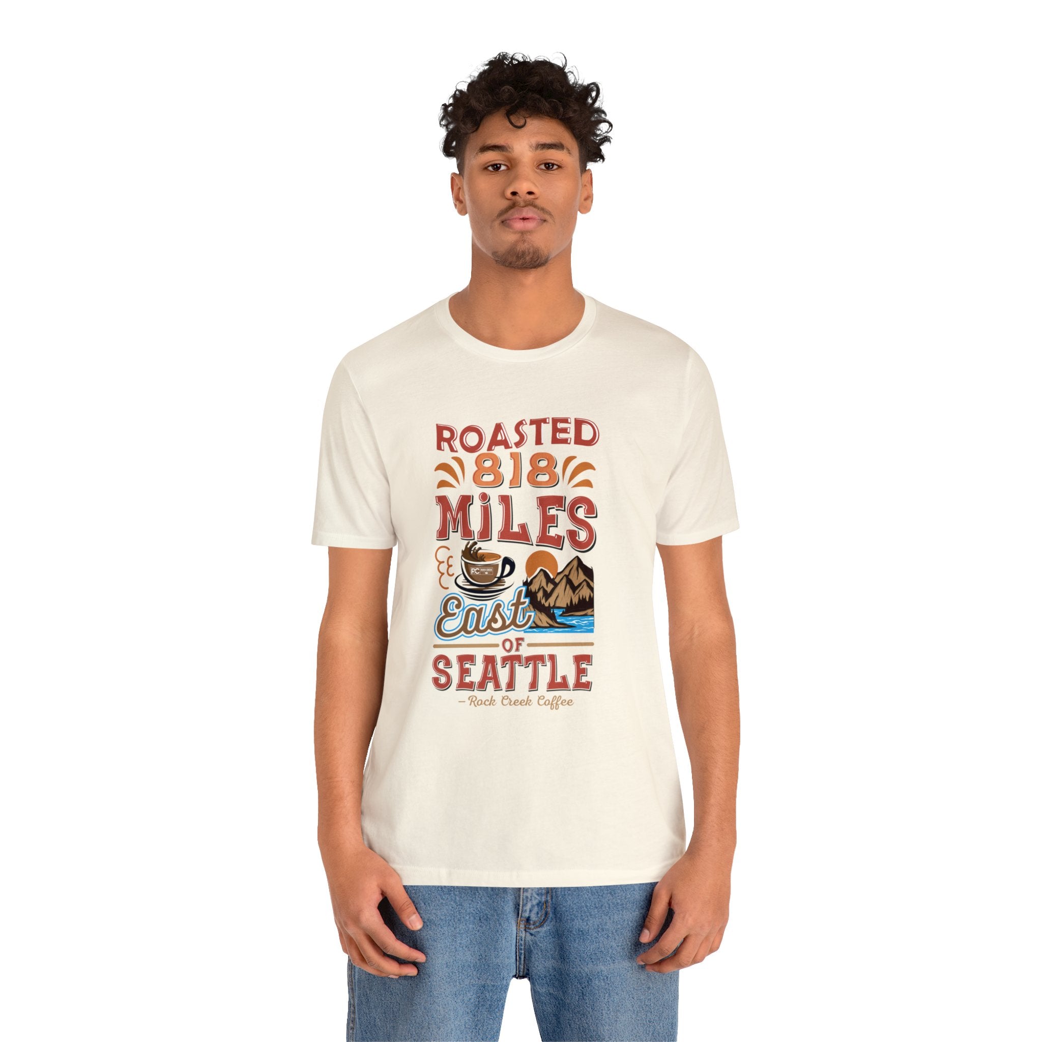 Adult man modeling a cream short-sleeve T-shirt printed with a colorful graphic that reads “Roasted 818 Miles East of Seattle,” featuring a steaming coffee cup and mountain lake illustration, shown against a plain white background.