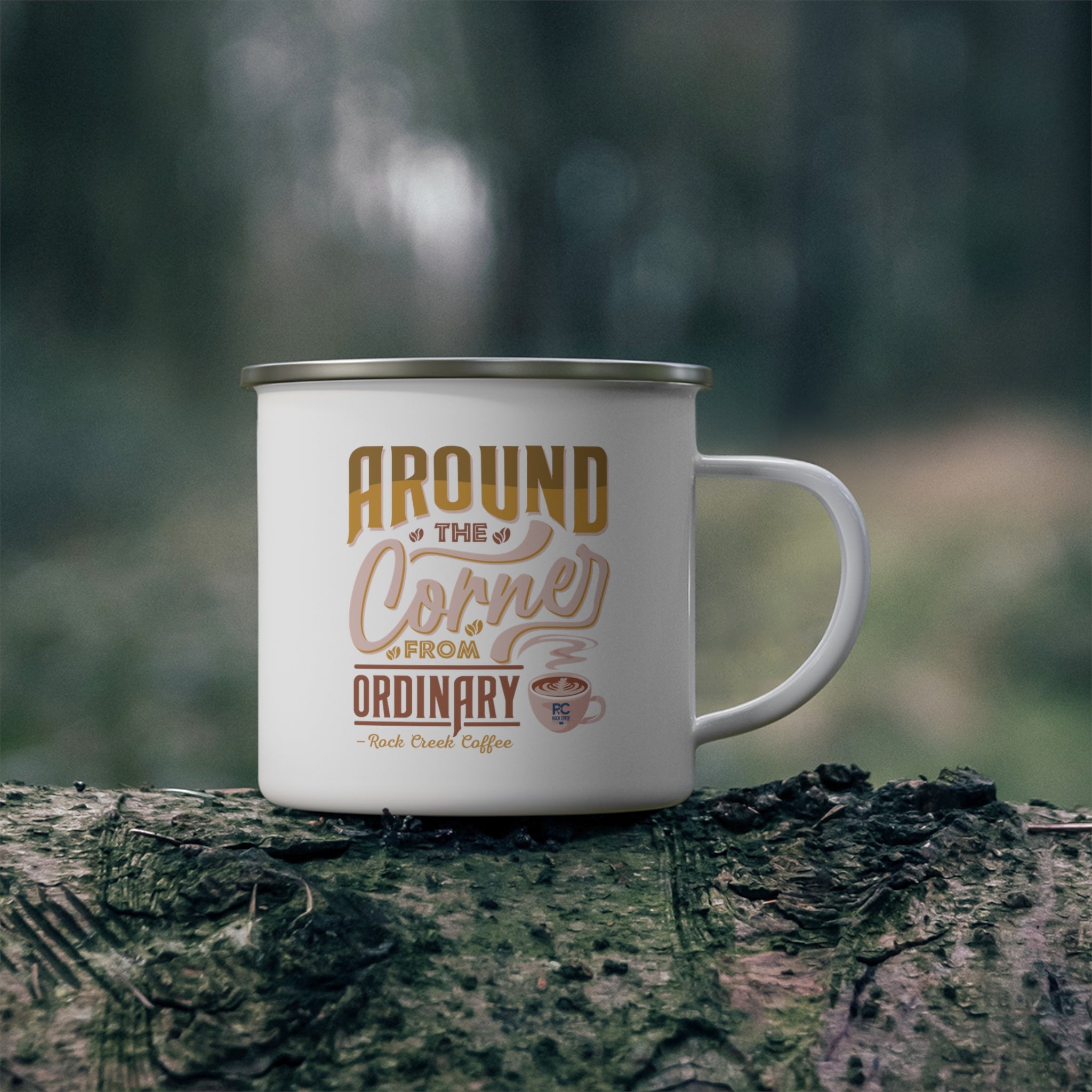 White enamel style coffee mug with silver rim and handle featuring the phrase Around the Corner from Ordinary Rock Creek Coffee in tan and brown lettering with a small latte cup illustration, placed on a mossy log outdoors with a blurred forest background.