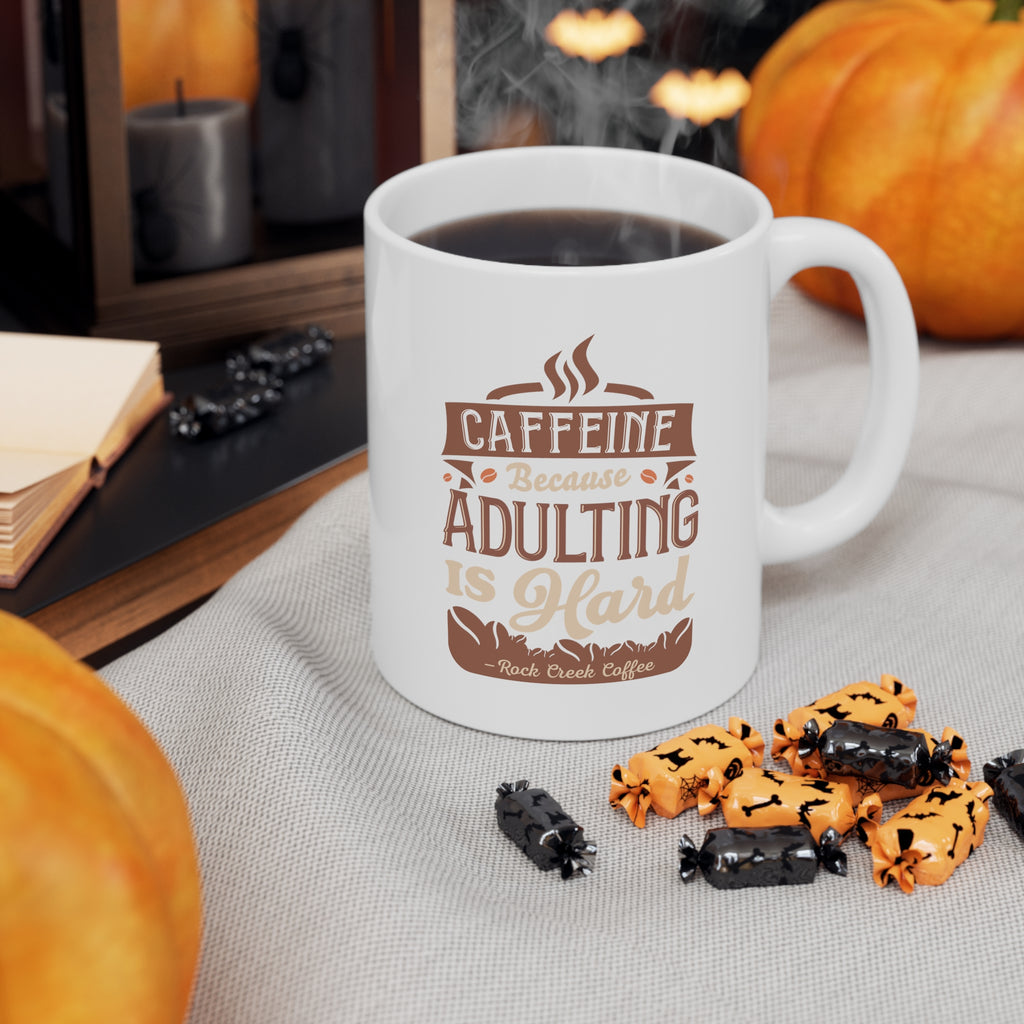 White Rock Creek Coffee mug featuring the phrase “Caffeine Because Adulting Is Hard” in brown and tan lettering, filled with coffee and styled with pumpkins and Halloween candies on a cozy fall themed tabletop.