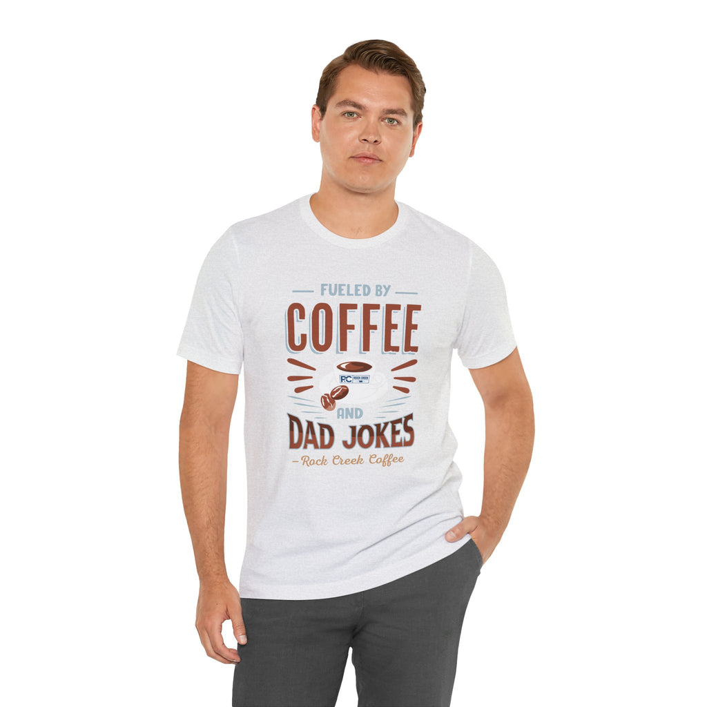 Adult man posing with one hand in his pocket while modeling an ash short-sleeve T-shirt featuring the “Fueled by Coffee and Dad Jokes” graphic with a coffee cup illustration and Rock Creek Coffee text, shown against a plain white background.