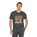 Adult man modeling an asphalt gray short-sleeve T-shirt featuring the “Around the Corner from Ordinary” graphic in a bold coffee-themed typographic design, posed with one hand in his pocket against a plain white background.