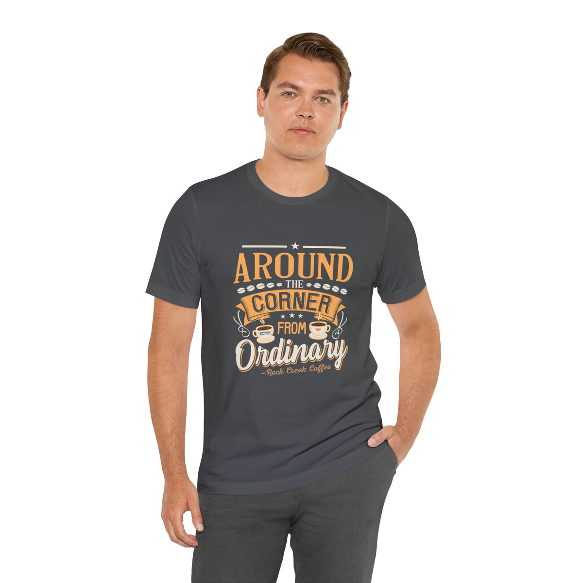 Adult man modeling an asphalt gray short-sleeve T-shirt featuring the “Around the Corner from Ordinary” graphic in a bold coffee-themed typographic design, posed with one hand in his pocket against a plain white background.