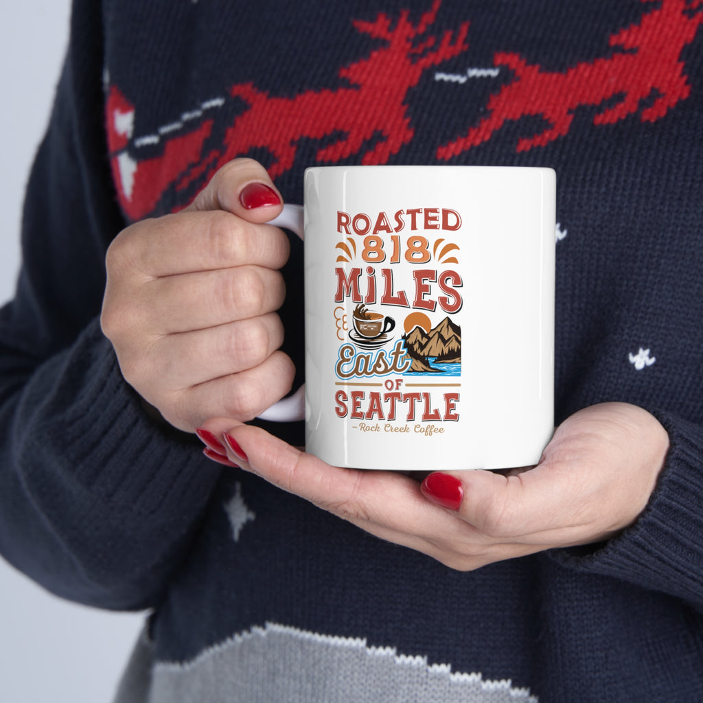Person wearing a navy holiday sweater holding a white Rock Creek Coffee mug featuring the colorful “Roasted 818 Miles East of Seattle” design.