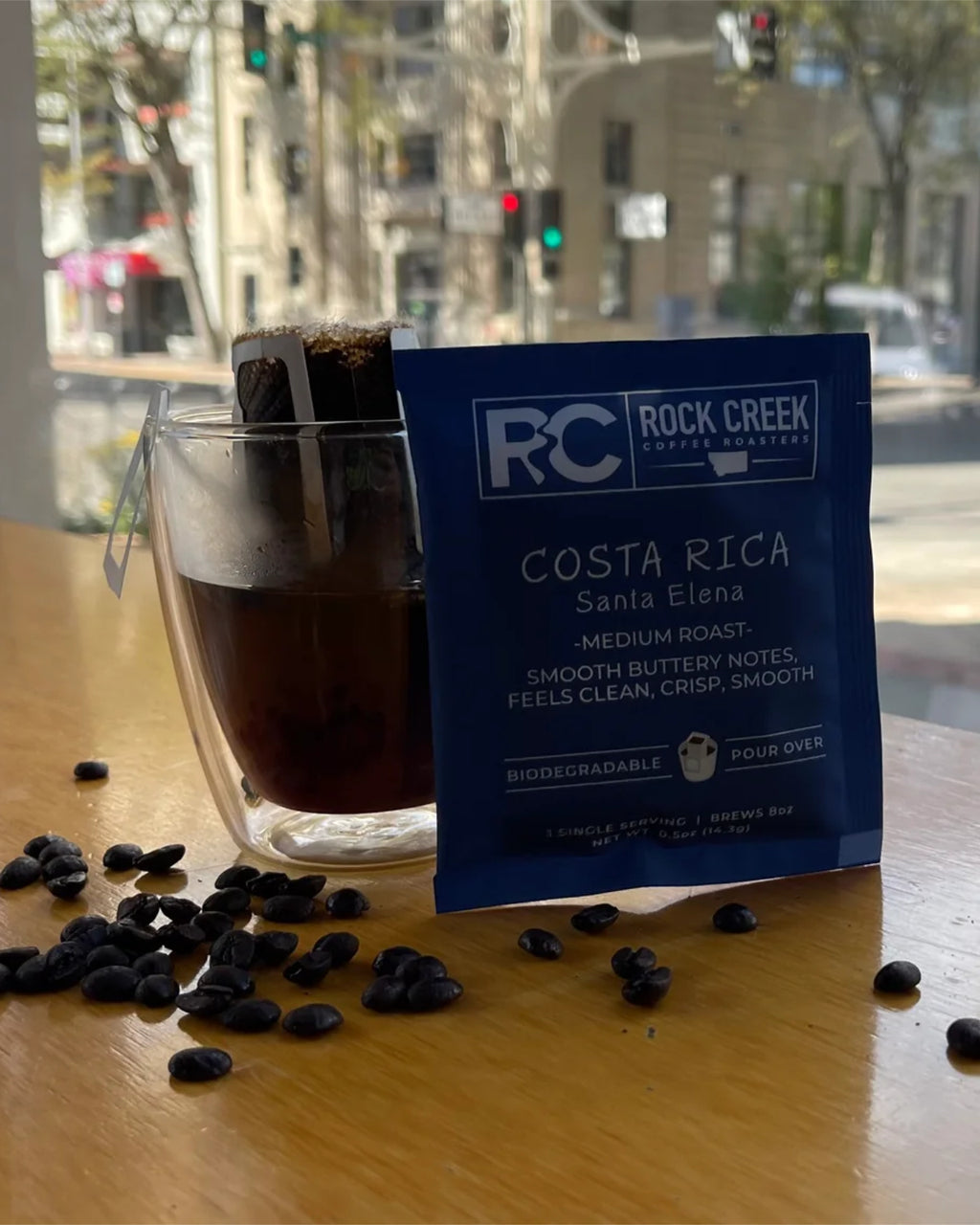 Rock Creek Coffee Roasters Costa Rica Santa Elena medium roast single-serve pour-over packet displayed beside a glass mug of freshly brewed coffee with a filter in place, coffee beans scattered on a wooden table, and a city street visible through a window in the background.