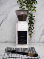 White Baratza Encore coffee grinder with beans in the hopper, set on a marble countertop with a striped cloth and cleaning brush in front, and green trailing plant against a marble wall background.
