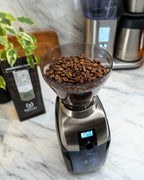 Baratza Virtuoso Plus coffee grinder with hopper full of whole coffee beans on a marble countertop, digital timer display set to 15.0 seconds, with a bag of coffee, houseplants, and a coffee maker in the background.