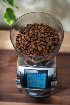 Top view of a Baratza Forte coffee grinder filled with whole coffee beans in a clear hopper, sitting on a wooden countertop with the digital touchscreen display visible below.