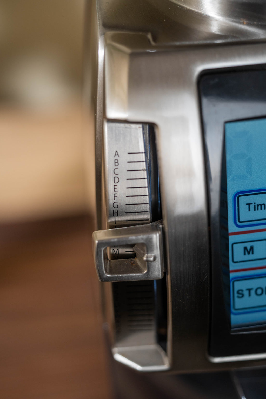 Close-up of the Baratza Forte grinder’s grind size adjustment lever with lettered scale markings (A–I) beside the touchscreen display, shown in brushed stainless steel.