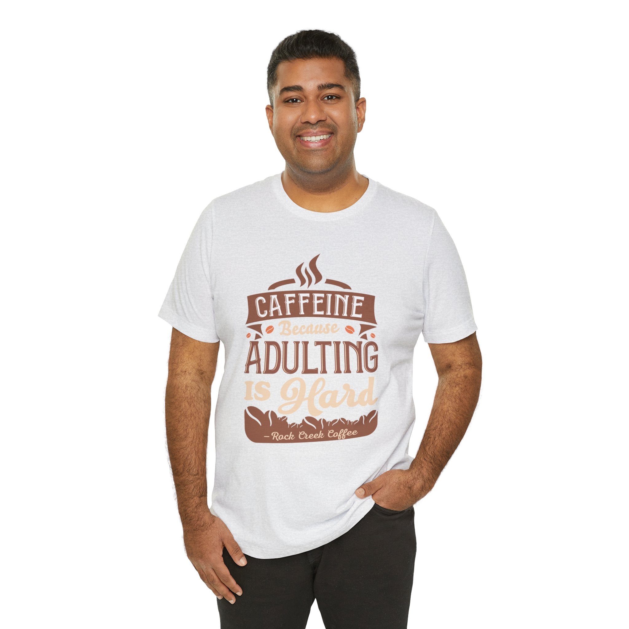 Adult man smiling while modeling an ash short-sleeve T-shirt printed with the phrase “Caffeine Because Adulting Is Hard” in a stacked typographic design, shown against a plain white background.