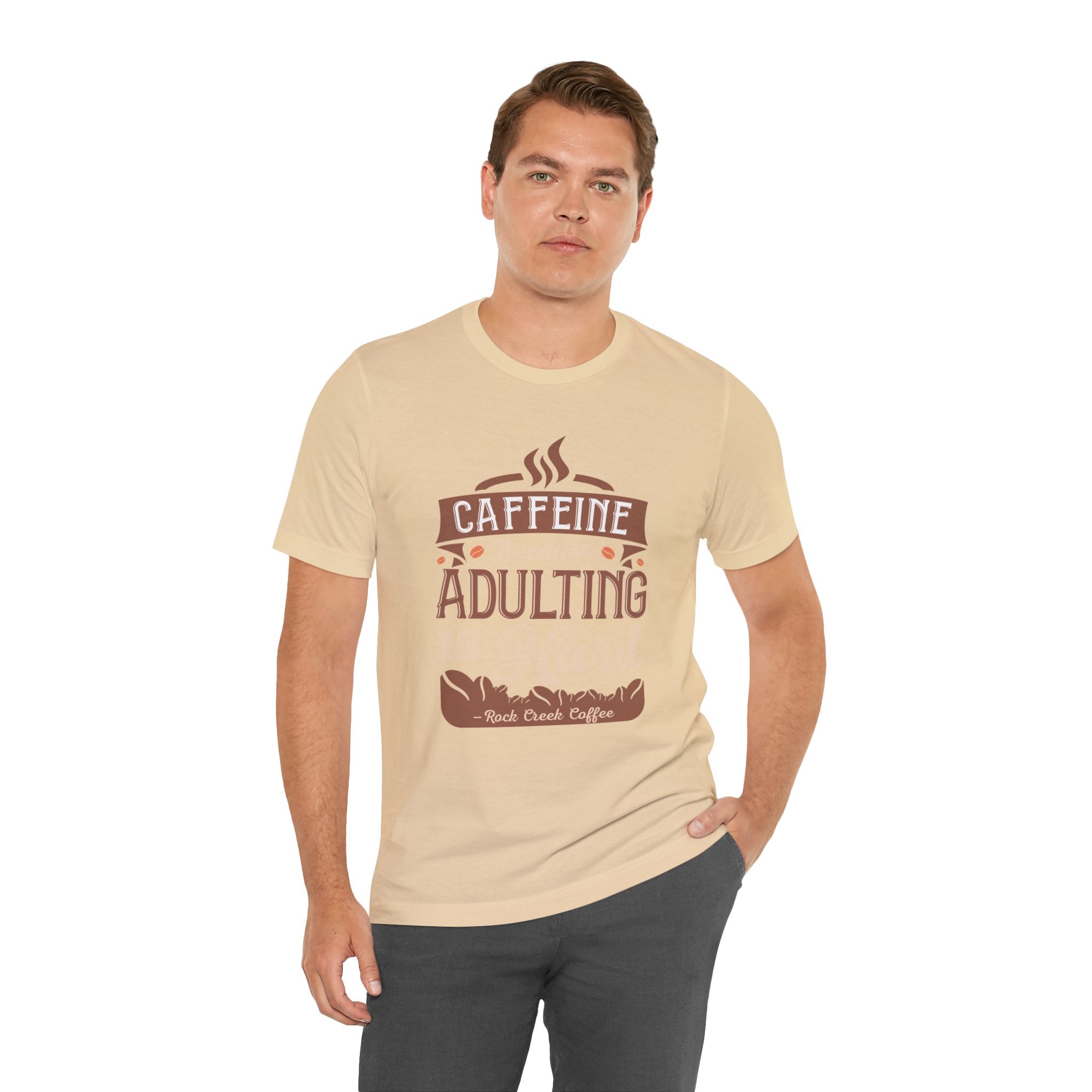 Adult man modeling a soft cream short-sleeve T-shirt featuring the “Caffeine Because Adulting Is Hard” graphic in a stacked typographic design, posed with one hand in his pocket against a plain white background.