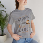 Adult woman modeling a heather gray short-sleeve T-shirt with the “Sippin on Genius Juice” graphic in bold stacked lettering and a small coffee cup illustration, photographed indoors beside a potted plant.