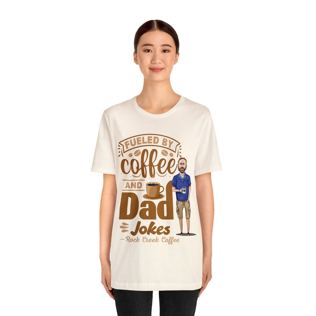 Woman wearing a cream graphic T-shirt with “Fueled by Coffee and Dad Jokes” design featuring coffee cup and cartoon dad.