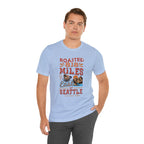 Adult man modeling a light blue short-sleeve T-shirt printed with a colorful graphic that reads “Roasted 818 Miles East of Seattle,” featuring a steaming coffee cup and mountain lake illustration, shown against a plain white background.