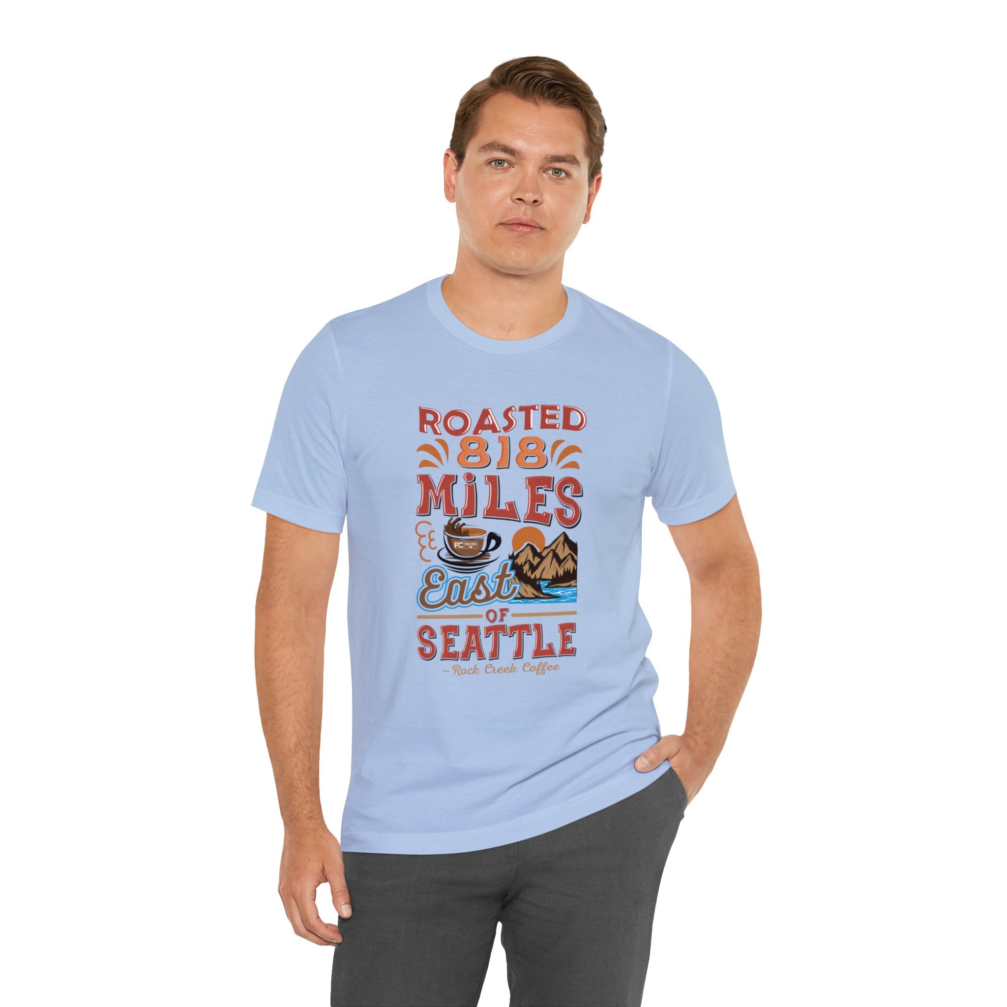 Adult man modeling a light blue short-sleeve T-shirt printed with a colorful graphic that reads “Roasted 818 Miles East of Seattle,” featuring a steaming coffee cup and mountain lake illustration, shown against a plain white background.