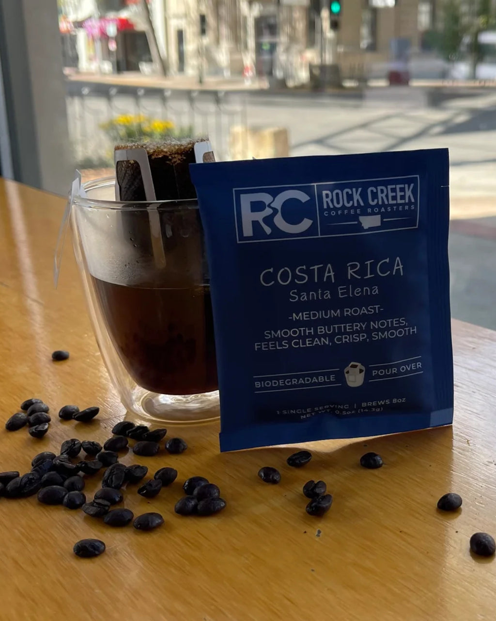 Rock Creek Coffee Roasters Costa Rica Santa Elena medium roast biodegradable single-serve pour-over packet displayed next to a glass mug of brewed coffee with the filter attached, coffee beans scattered on a wooden table, and a city street scene visible through a window in the background.