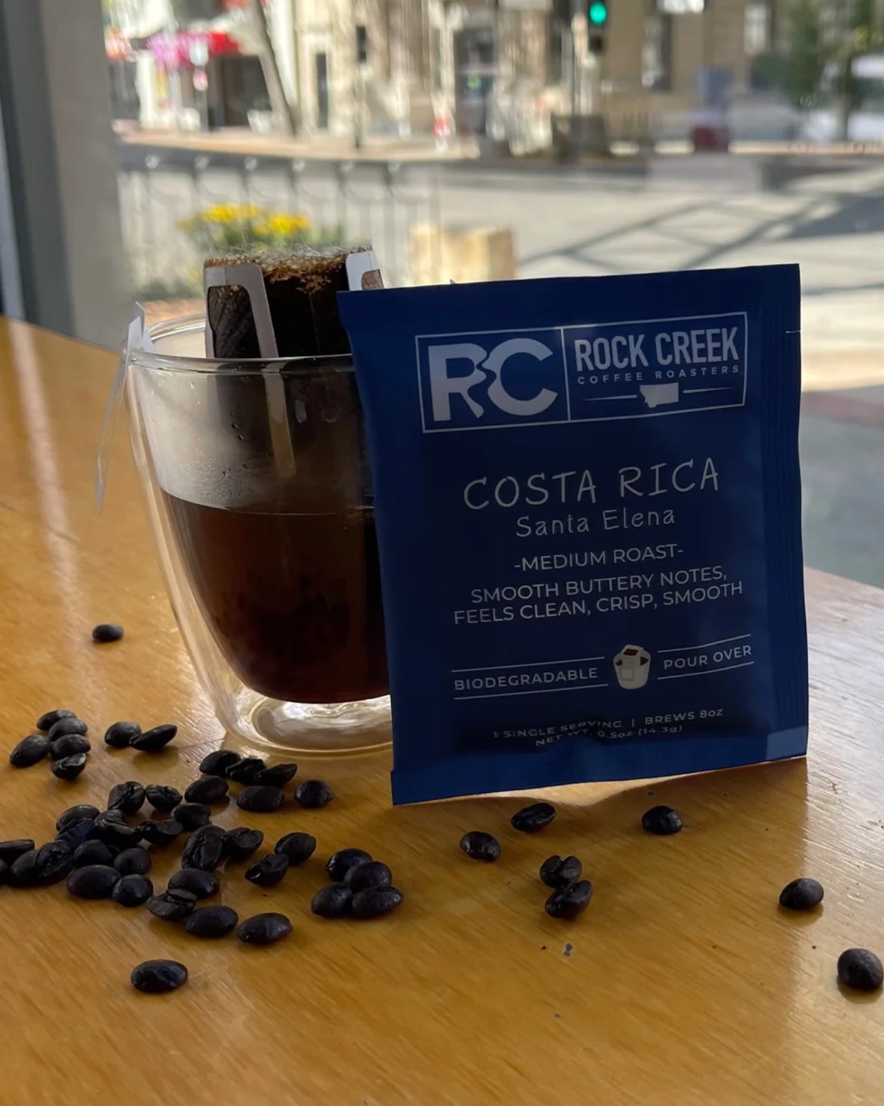Rock Creek Coffee Roasters Costa Rica Santa Elena medium roast biodegradable single-serve pour-over packet displayed next to a glass mug of brewed coffee with the filter attached, coffee beans scattered on a wooden table, and a city street scene visible through a window in the background.