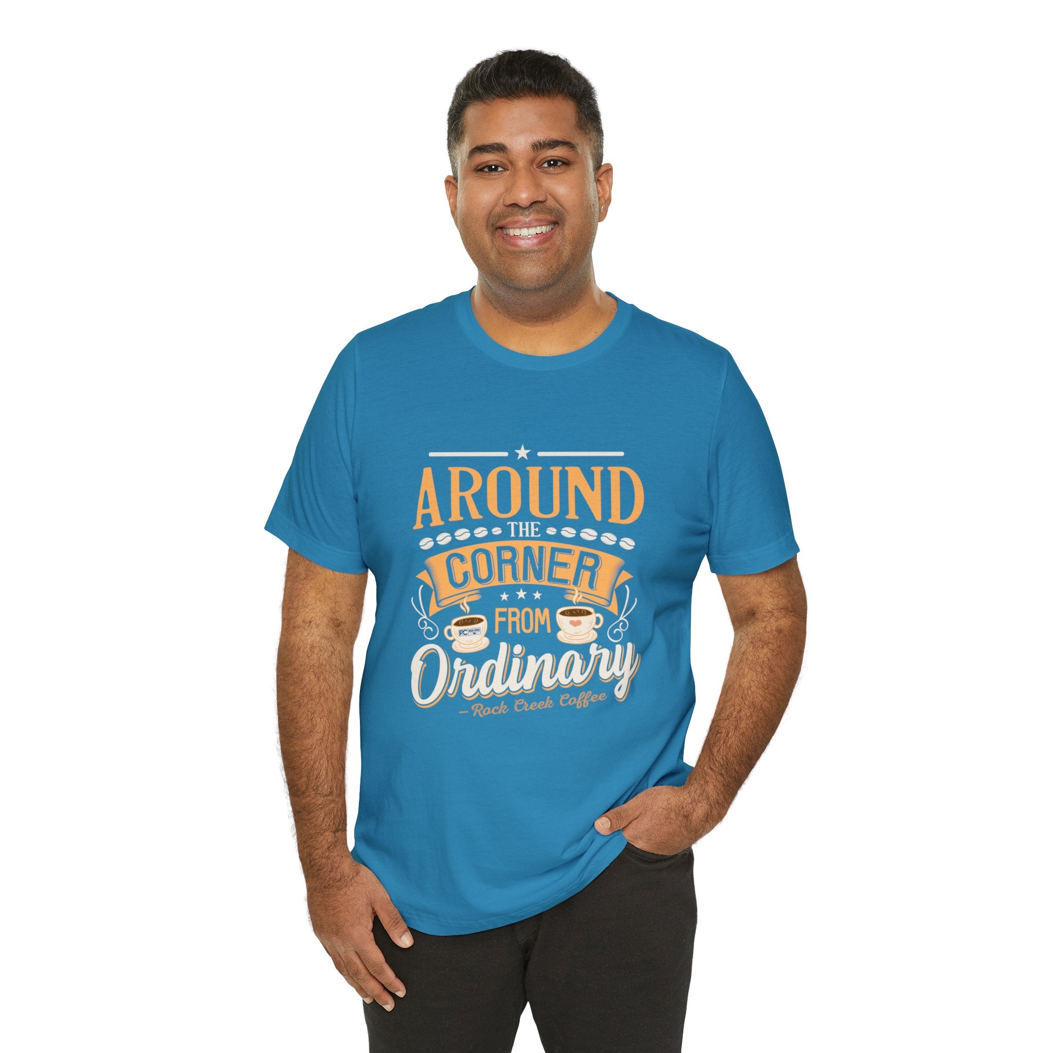 Adult man smiling while modeling an aqua short-sleeve T-shirt featuring the phrase “Around the Corner from Ordinary” in a bold typographic coffee-themed design, shown against a plain white background.