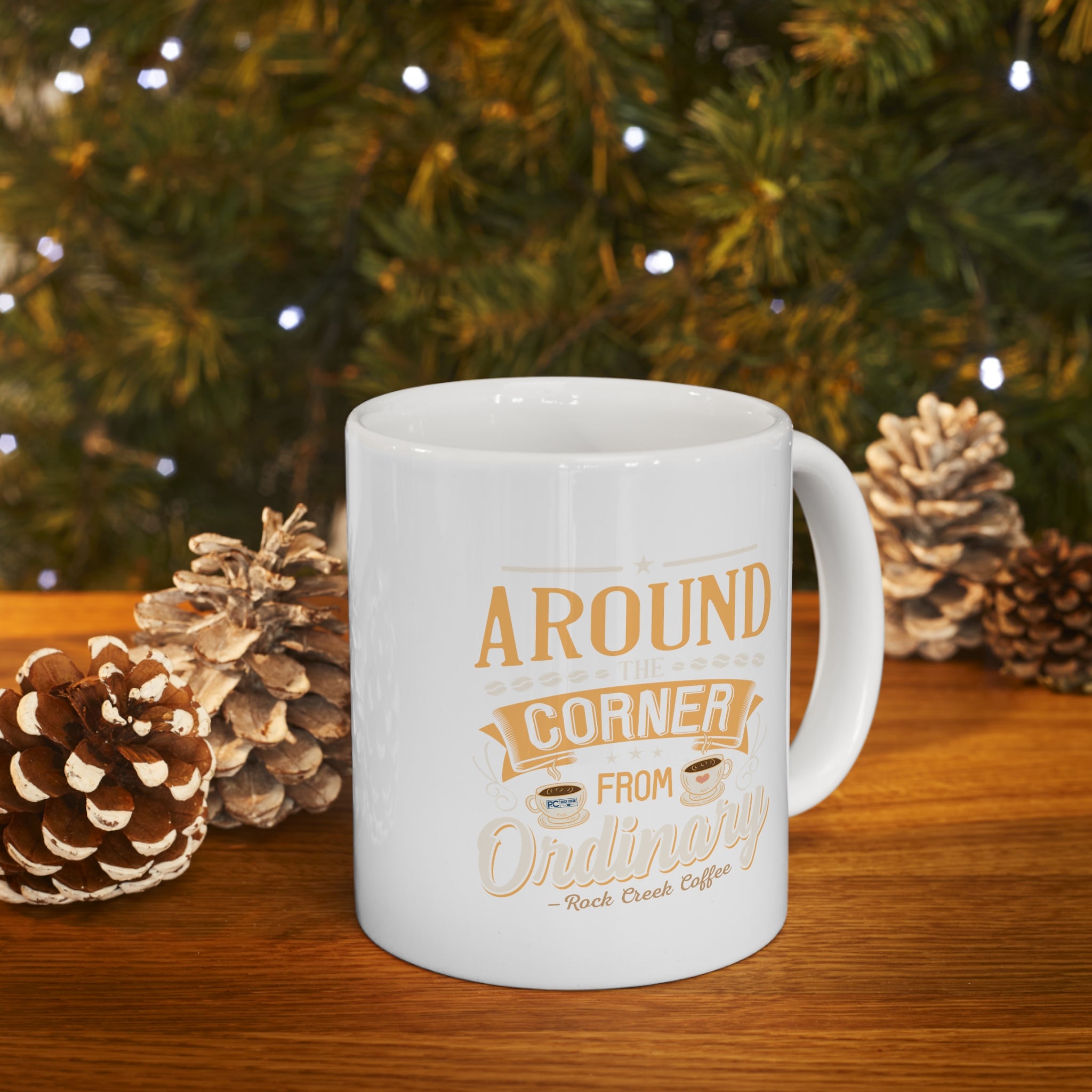 White Rock Creek Coffee mug with “Around the Corner from Ordinary” design in gold and cream lettering displayed on a wooden table with pinecones and soft holiday lights in the background.