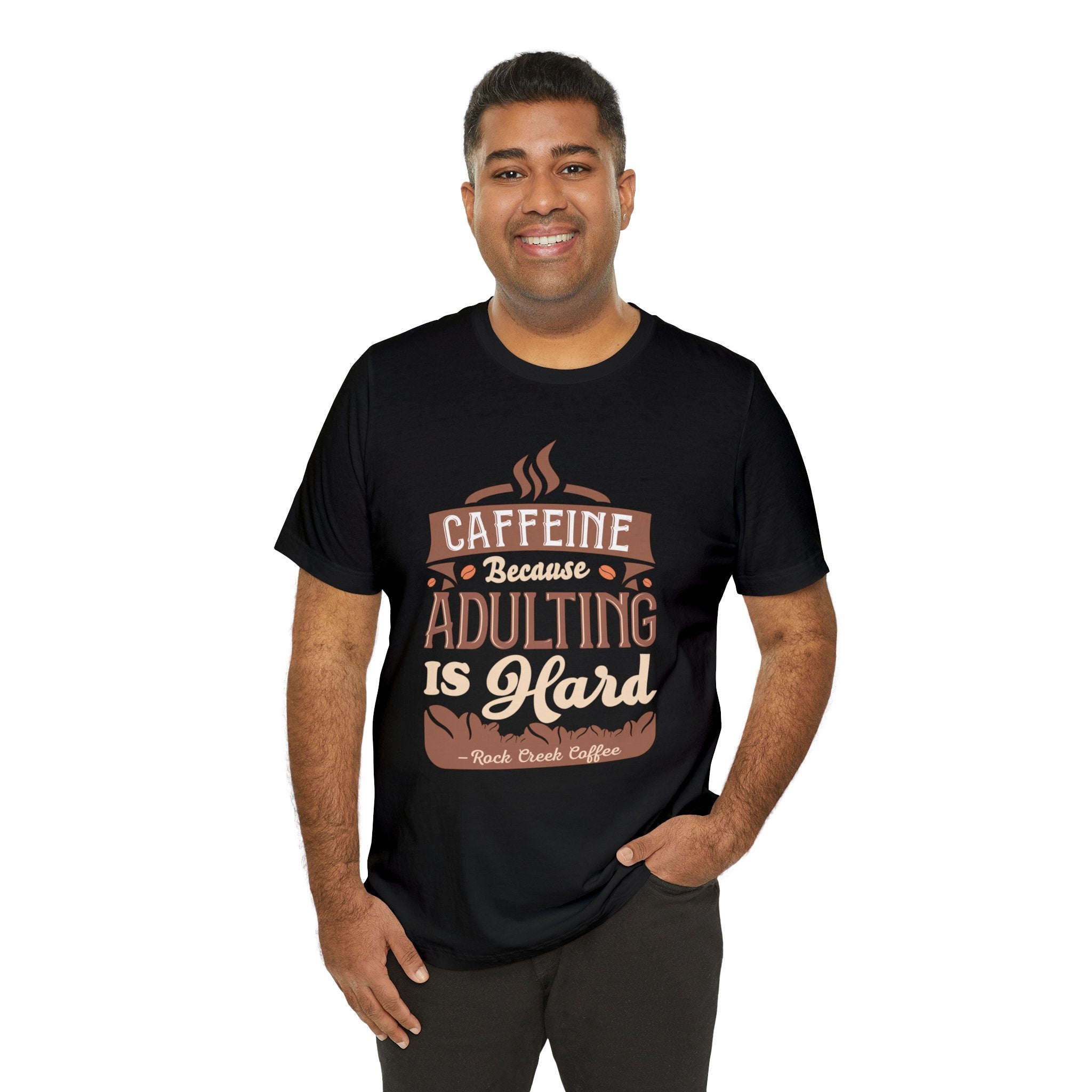 Adult man smiling while modeling a black short-sleeve T-shirt printed with the phrase “Caffeine Because Adulting Is Hard” in a stacked typographic design, shown against a plain white background.