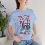 Adult woman modeling a light blue short-sleeve Seattle coffee graphic T-shirt reading “Roasted 818 Miles East of Seattle,” shown against a plain white background with a potted plant nearby.