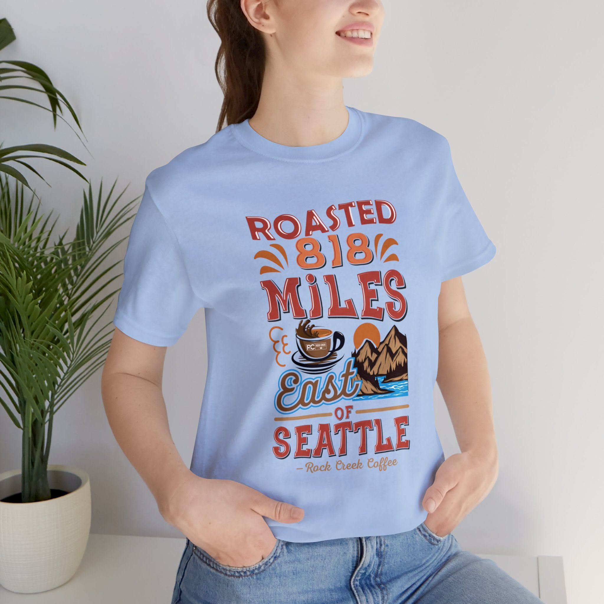 Adult woman modeling a light blue short-sleeve Seattle coffee graphic T-shirt reading “Roasted 818 Miles East of Seattle,” shown against a plain white background with a potted plant nearby.