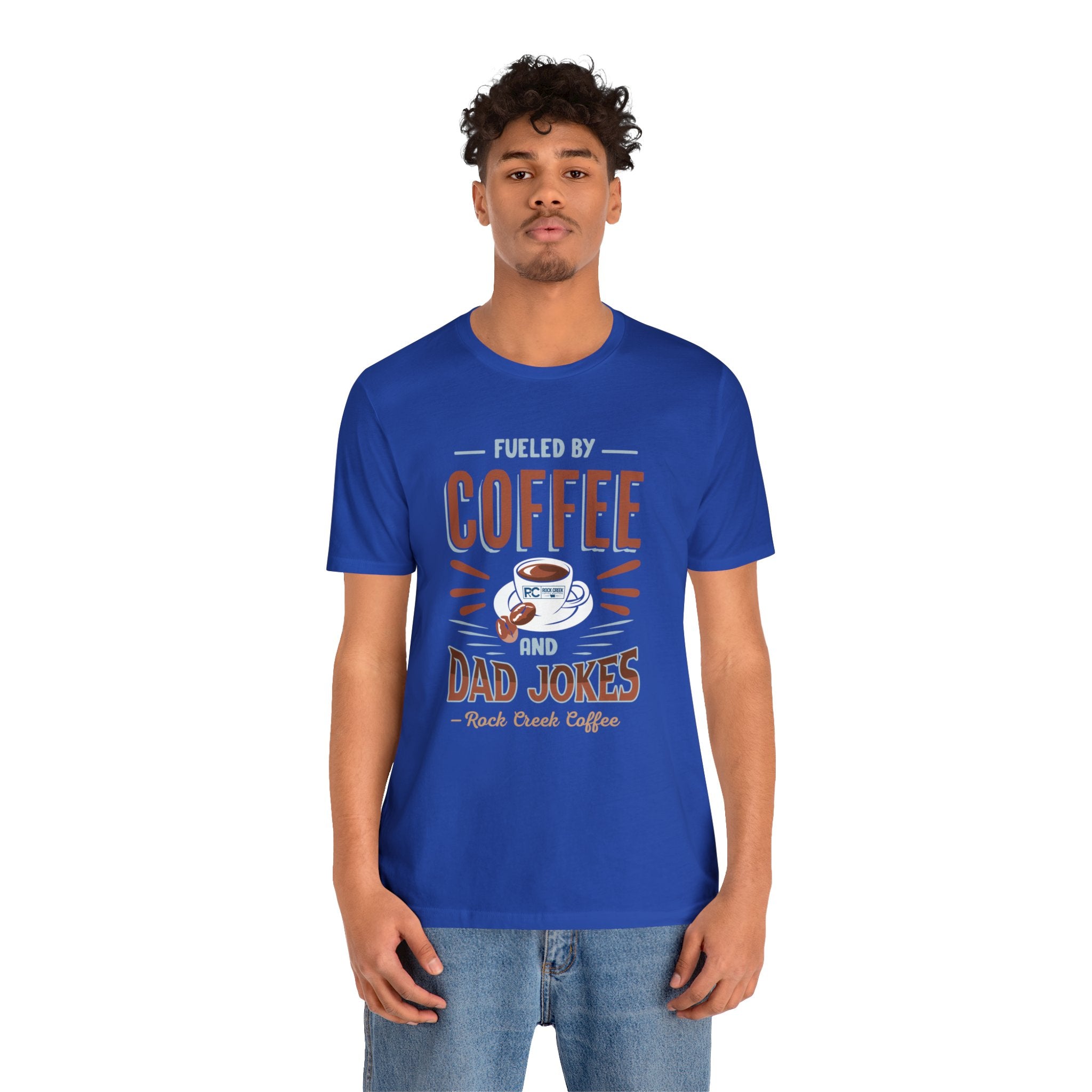 Adult man modeling a royal blue short-sleeve T-shirt featuring the “Fueled by Coffee and Dad Jokes” graphic with a coffee cup illustration and Rock Creek Coffee text, shown against a plain white background.