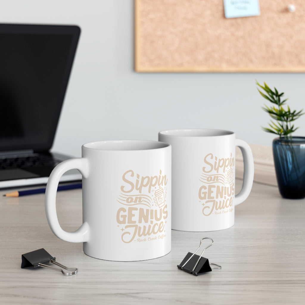 Two white Rock Creek Coffee mugs featuring the phrase “Sippin on Genius Juice” in beige lettering with an RC coffee cup graphic, displayed on a desk with a laptop, binder clips, books, and a small potted plant in the background.