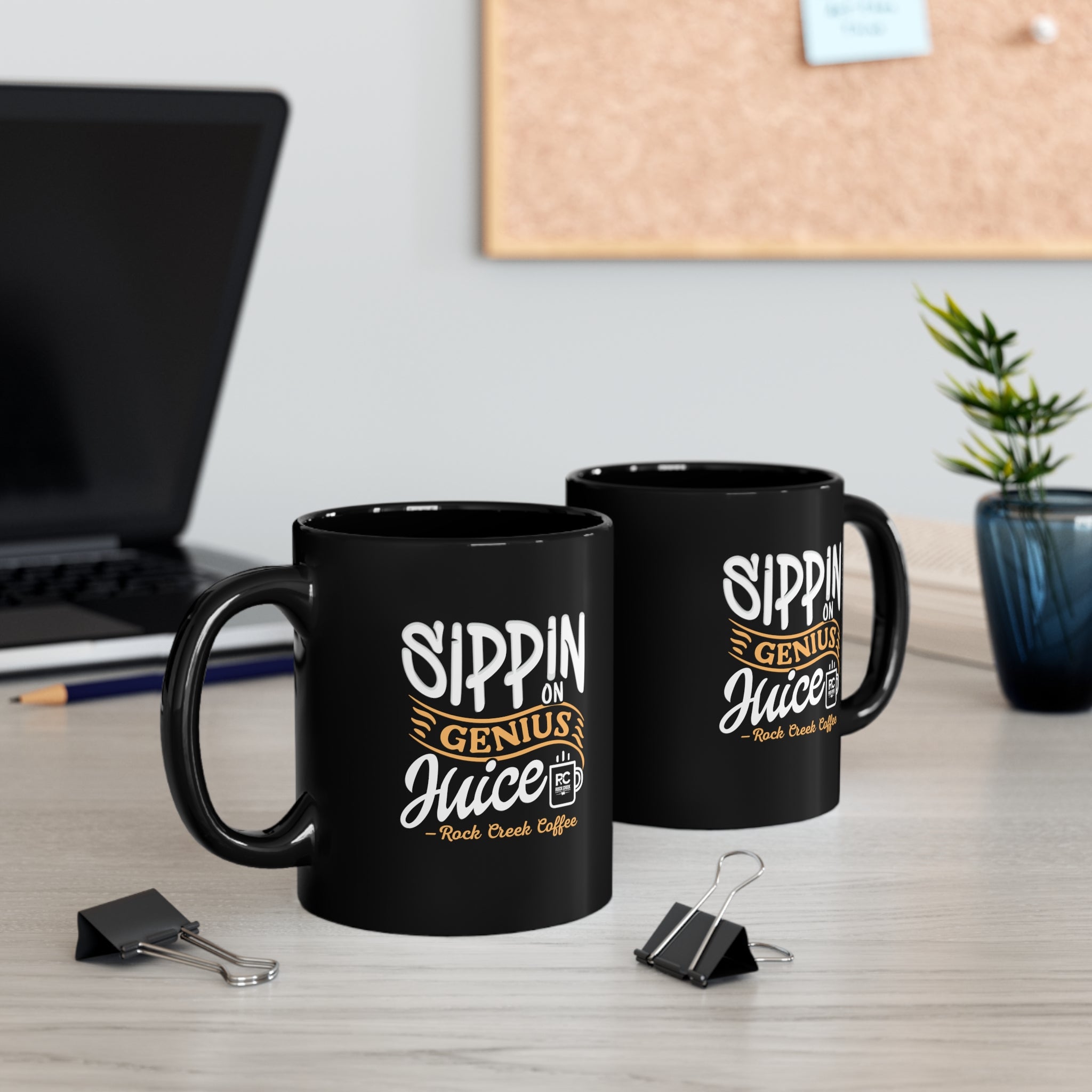 Two black ceramic coffee mugs featuring the phrase “Sippin on Genius Juice Rock Creek Coffee” in white and gold lettering, displayed on a desk with a laptop, binder clips, and a small potted plant in the background.