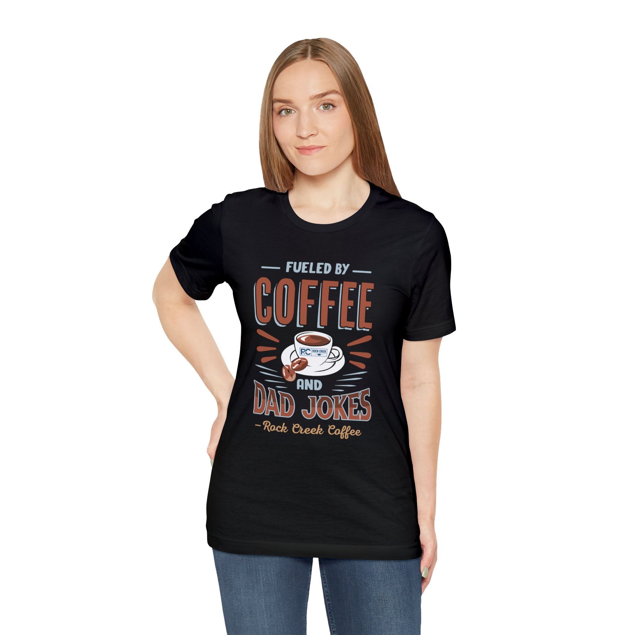 Adult woman modeling a black short-sleeve T-shirt featuring the “Fueled by Coffee and Dad Jokes” graphic with a coffee cup illustration and Rock Creek Coffee text, shown against a plain white background.