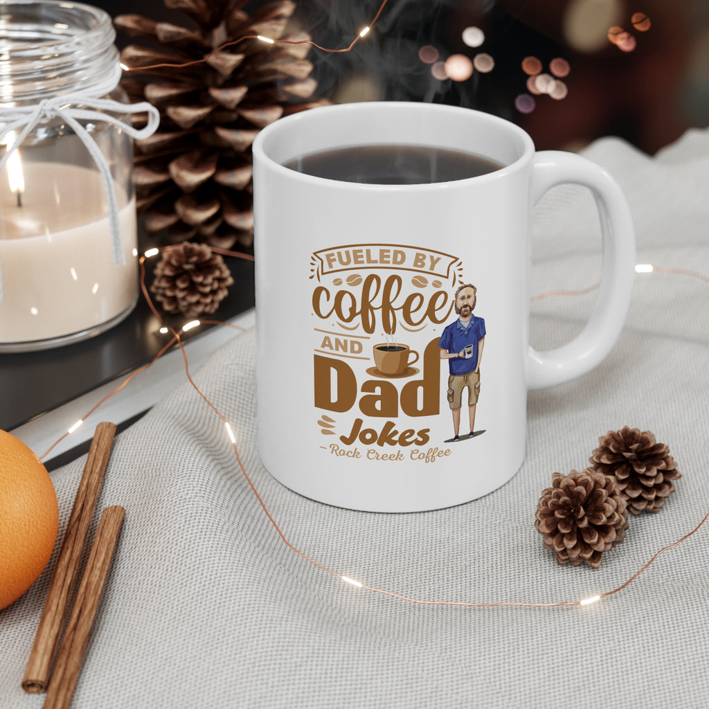 White ceramic coffee mug filled with coffee featuring the text Fueled by Coffee and Dad Jokes Rock Creek Coffee with a cartoon illustration of a man holding a coffee cup, styled with pinecones, a candle, cinnamon sticks, and string lights.