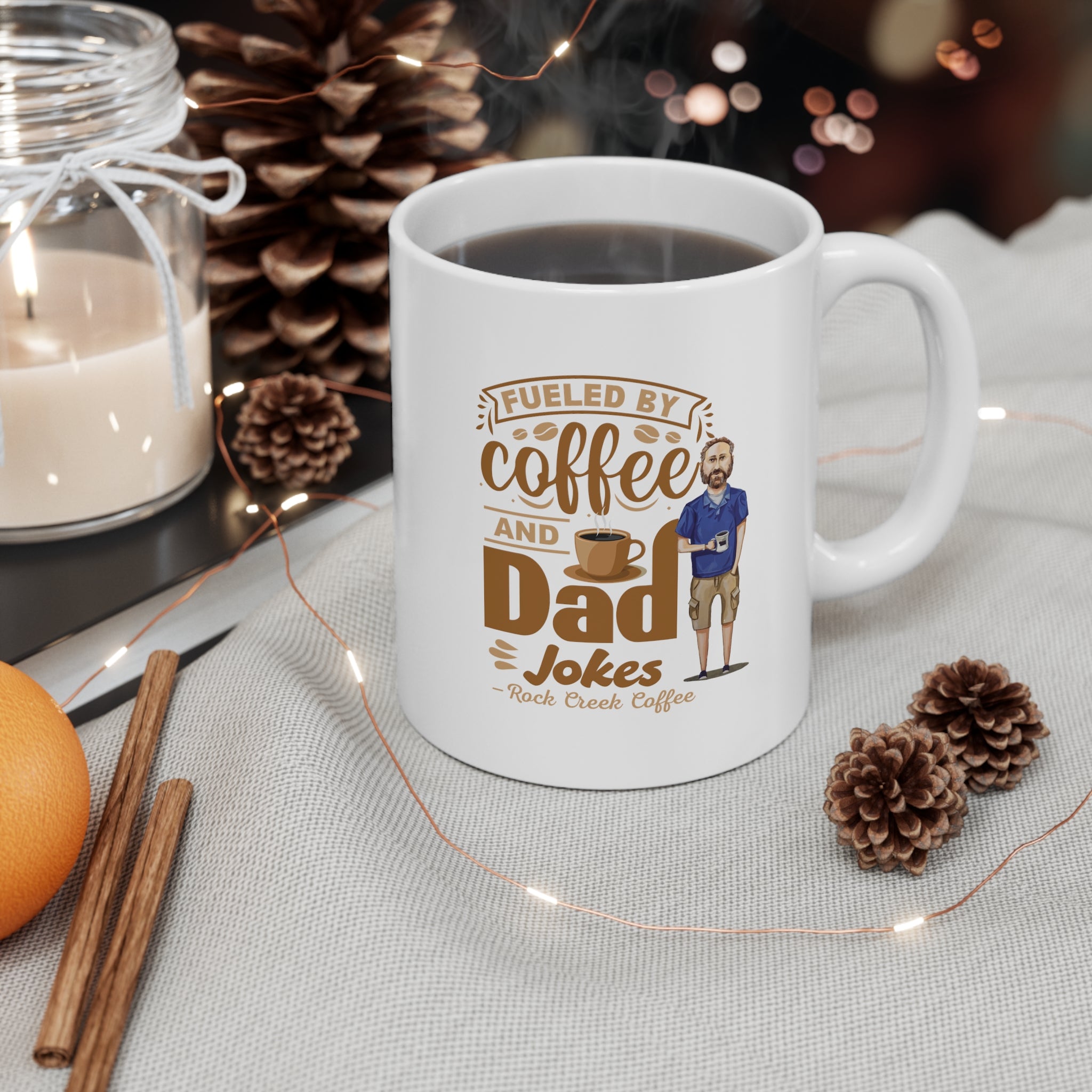 White ceramic coffee mug filled with coffee featuring the text Fueled by Coffee and Dad Jokes Rock Creek Coffee with a cartoon illustration of a man holding a coffee cup, styled with pinecones, a candle, cinnamon sticks, and string lights.