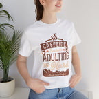 Adult woman modeling a white short-sleeve T-shirt featuring the “Caffeine Because Adulting Is Hard” graphic in a stacked typographic design, photographed indoors beside a potted plant against a light neutral background.