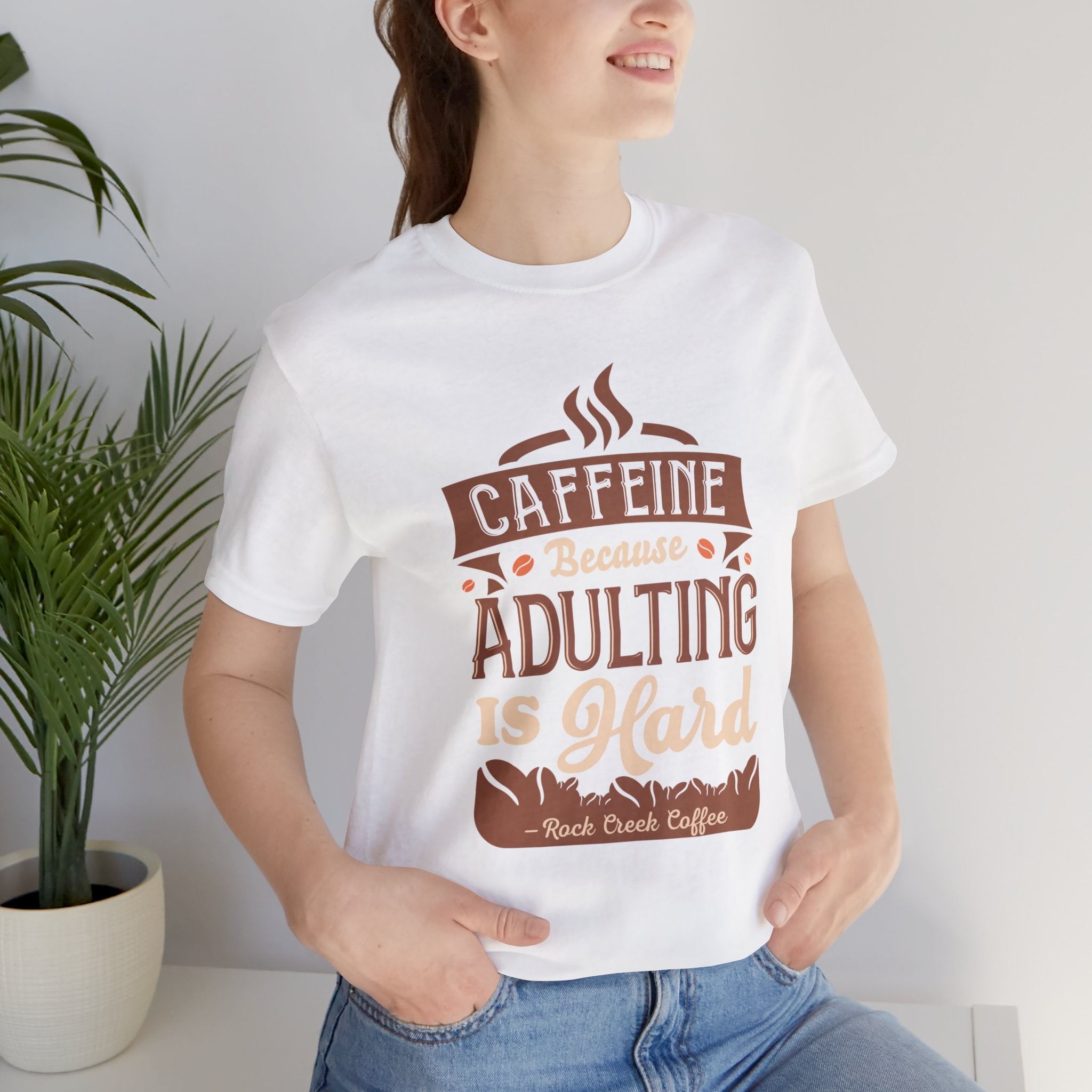 Adult woman modeling a white short-sleeve T-shirt featuring the “Caffeine Because Adulting Is Hard” graphic in a stacked typographic design, photographed indoors beside a potted plant against a light neutral background.