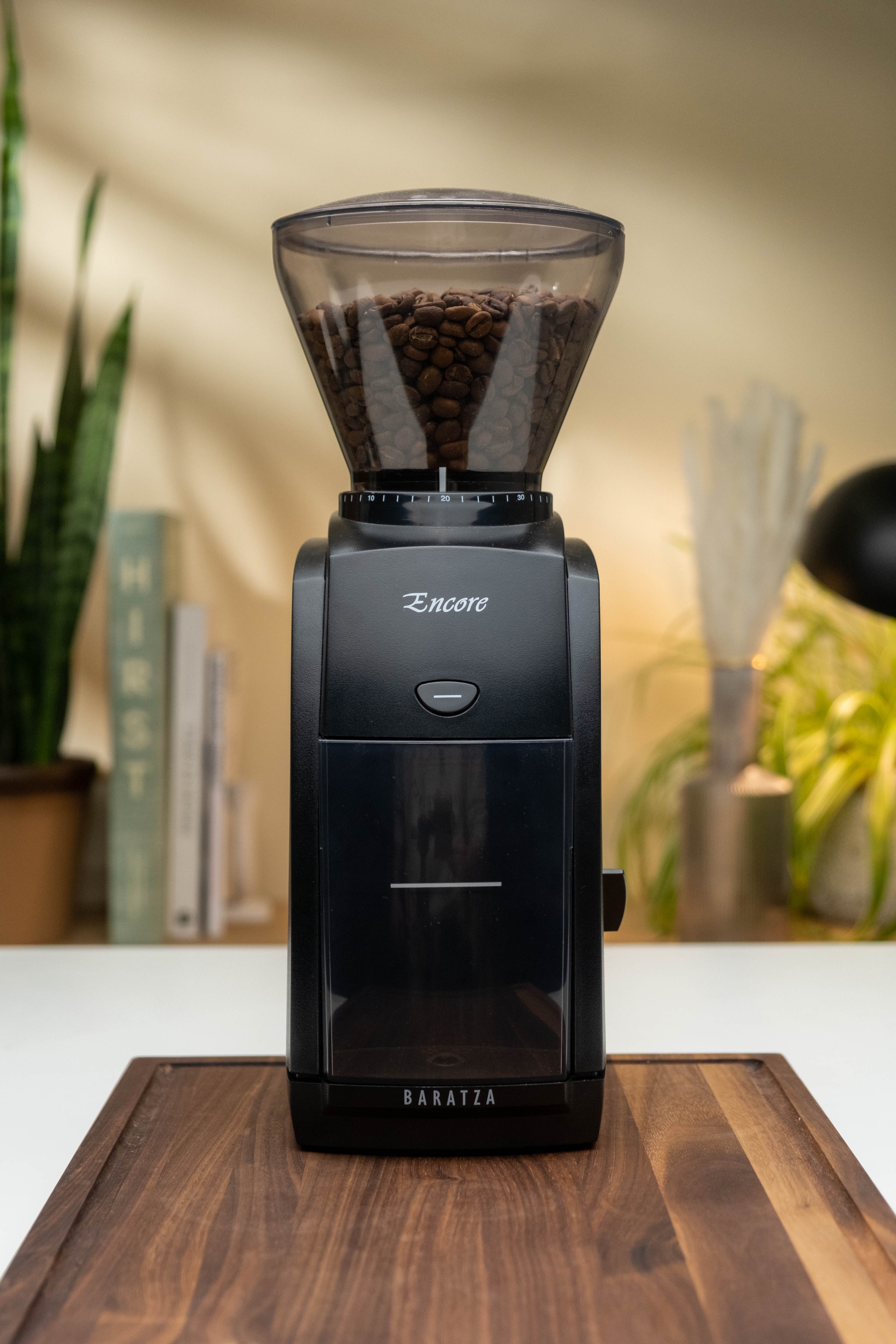 Black Baratza Encore coffee grinder with beans in the hopper, placed on a wooden board in a home kitchen setting with plants and books in the background.