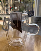 Single-serve pour-over coffee filter filled with ground coffee hanging over a clear double-walled glass mug on a wooden table, with a city street visible through a window in the background.