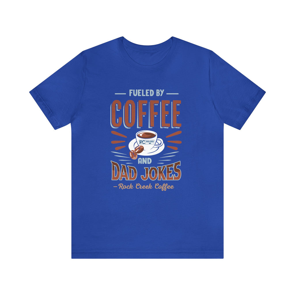 Royal blue short-sleeve T-shirt displayed flat against a plain white background, featuring a bold “Fueled by Coffee and Dad Jokes” graphic with a coffee cup illustration and Rock Creek Coffee text below.