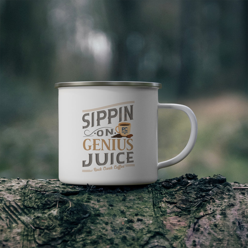 White enamel-style coffee mug with silver rim and handle featuring the phrase “Sippin on Genius Juice - Rock Creek Coffee” in gray and tan lettering with a small coffee cup illustration, placed on a mossy log outdoors with a blurred forest background.