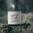 White enamel-style coffee mug with silver rim and handle featuring the phrase “Sippin on Genius Juice - Rock Creek Coffee” in gray and tan lettering with a small coffee cup illustration, placed on a mossy log outdoors with a blurred forest background.