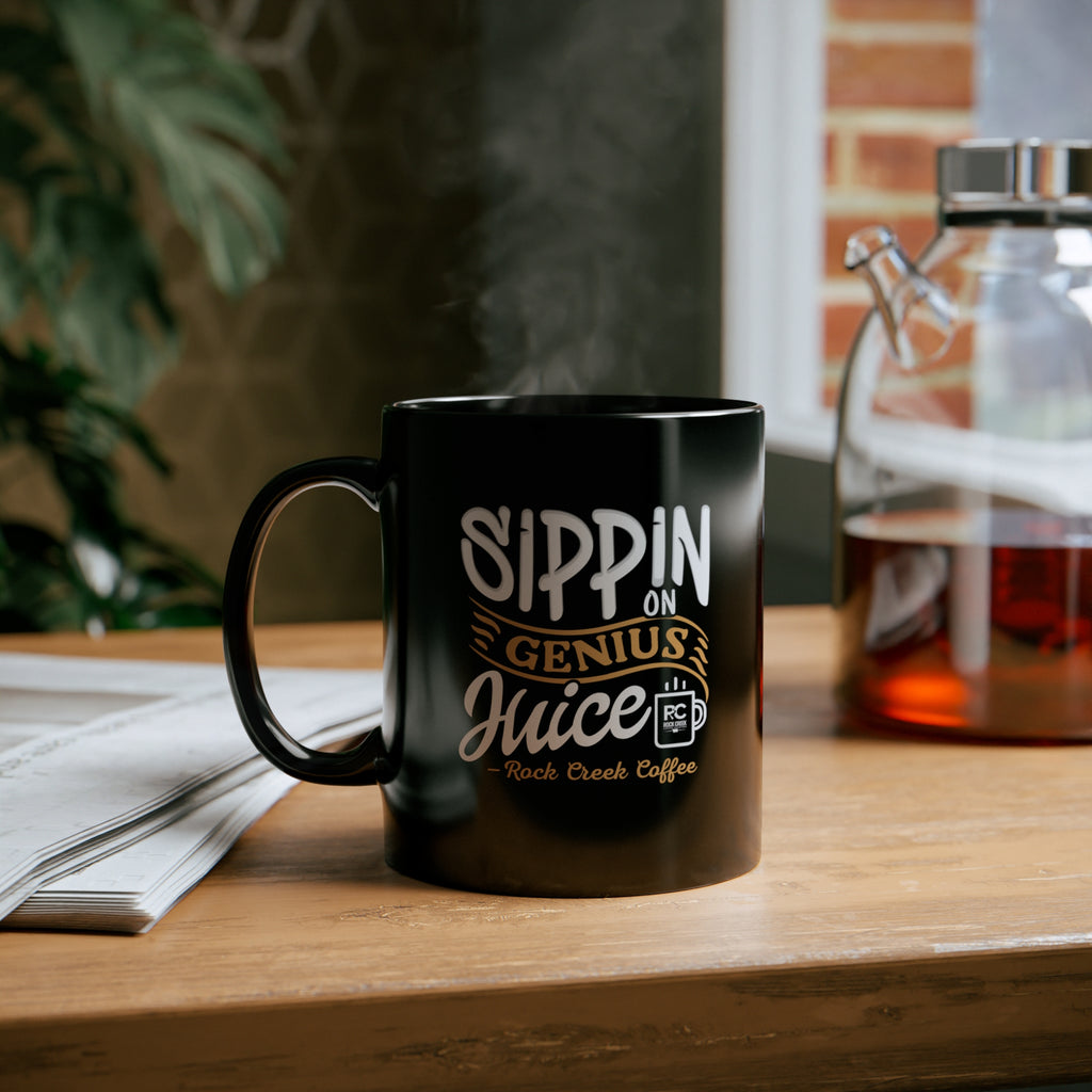 Black ceramic coffee mug with “Sippin on Genius Juice Rock Creek Coffee” design in white and gold lettering, filled with hot coffee and steam rising, placed on a wooden desk next to an open newspaper with a glass coffee carafe in the background.
