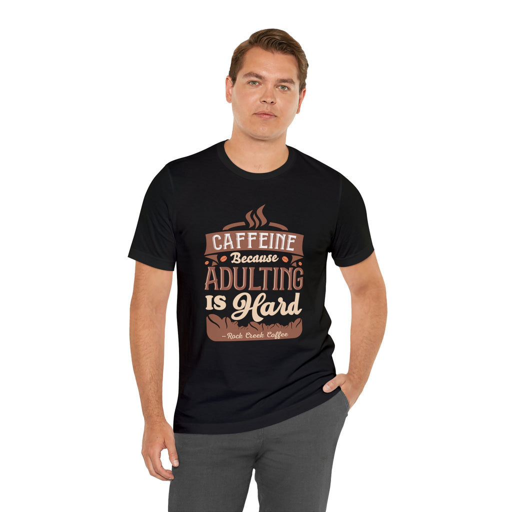 Adult man posing with one hand in his pocket while modeling a black short-sleeve T-shirt featuring the “Caffeine Because Adulting Is Hard” graphic in a stacked typographic design, shown against a plain white background.