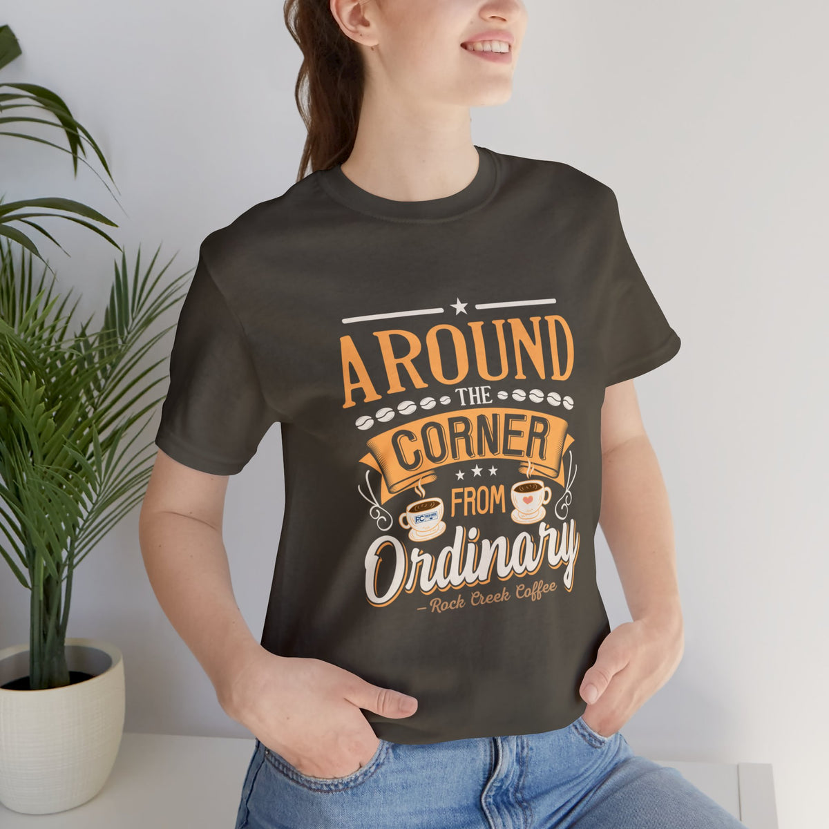 Adult woman modeling an army green short-sleeve T-shirt featuring the “Around the Corner from Ordinary” graphic in a bold coffee-themed typographic design, photographed indoors beside a potted plant against a light neutral background.