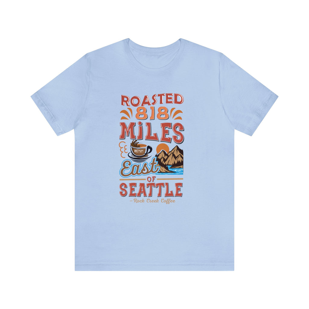 Light blue short-sleeve T-shirt displayed flat on a white background featuring a colorful graphic that reads “Roasted 818 Miles East of Seattle,” with a steaming coffee cup and mountain lake illustration on the front.